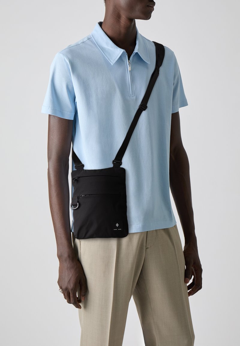 Black crossbody bag made of fabric, featuring multiple zip pockets and a shoulder strap. Worn over a light blue polo and beige trousers.