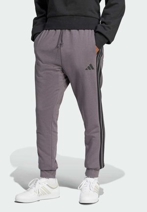 Pantalones deportivos grises con cintura elástica y puños acanalados, con franjas negras a los lados y un logo de Adidas en el muslo izquierdo. Combinados con zapatillas blancas.