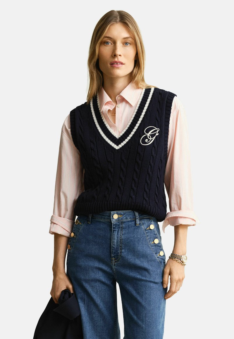 Femme portant un gilet sans manches en maille torsadée bleu marine avec bordure blanche, par-dessus une chemise rose à col, et un jean bleu taille haute avec des boutons dorés.