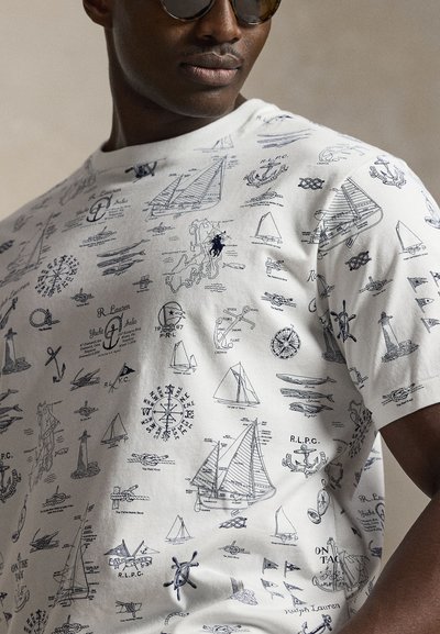 Άνδρας που φοράει λευκό T-shirt με μαύρες ναυτικές εικόνες, όπως ιστιοφόρα, άγκυρες, πυξίδες και λεπτομέρειες φάρου.