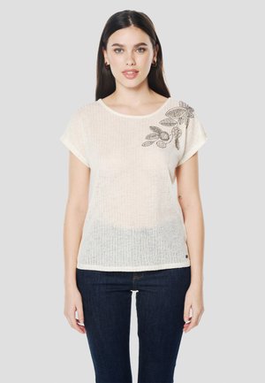 Lekka, kremowa bluzka o luźnym kroju, z okrągłym dekoltem, krótkimi rękawami i koralikowym kwiatowym wzorem na lewym ramieniu.