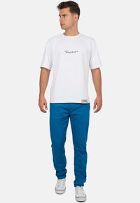 Witte katoenen t-shirt met zwart logo op de borst, gecombineerd met blauwe broek en witte sneakers. Casual pasvorm en eenvoudig ontwerp met korte mouwen.