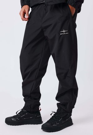 Personne portant un pantalon de sport noir avec le logo PXP et des baskets noires, debout les mains dans les poches devant un fond uni.