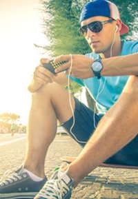 Un giovane uomo è seduto su uno skateboard, indossa occhiali da sole, una camicia blu e pantaloni corti. Tiene un dispositivo con degli auricolari collegati.