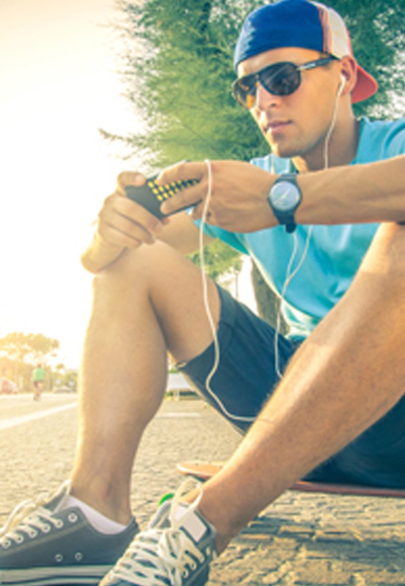 Un giovane uomo è seduto su uno skateboard, indossa occhiali da sole, una camicia blu e pantaloni corti. Tiene un dispositivo con degli auricolari collegati.