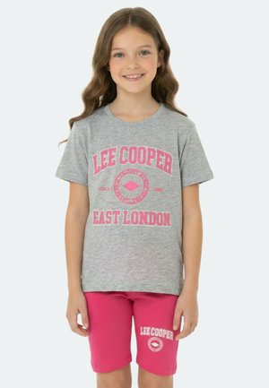 Jeune fille souriante aux cheveux longs et ondulés, portant un t-shirt gris Lee Cooper East London et un short rose, sur un fond uni.