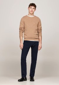 Maglione di lana marrone con polsini a costine, indossato sopra una t-shirt bianca; abbinato a pantaloni blu scuro e scarpe nere lucide.