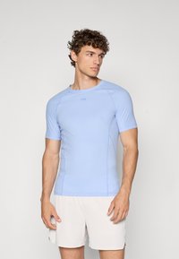 Lyseblå treningstrøye med korte ermer, tettsittende design og teksturerte paneler i sidene. Brukt sammen med lyse shorts.
