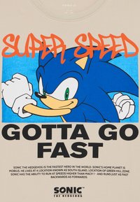 Bežinė medvilninė marškinė su grafiniu atvaizdu mėlynos ežiuko su žaliomis akimis, ryškiai oranžine tekstu "SUPER SPEED" ir juodu tekstu "GOTTA GO FAST."