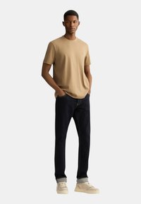 T-shirt beige a girocollo, jeans in denim scuro e sneakers di colore chiaro. Il modello è in piedi con le mani in tasca, mostrando il design dell'outfit casual.