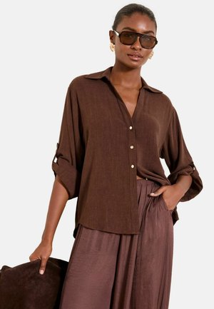 Femeie purtând o cămașă maro supradimensionată cu nasturi, mânecile suflecate, pantaloni largi asortați, ochelari de soare mari și cercei aurii, cu mâna în buzunar.