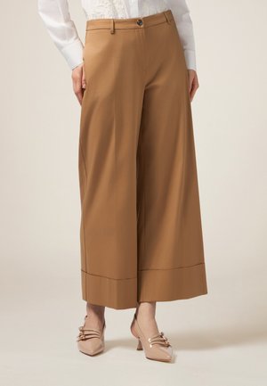 Pantaloni beige a gamba larga con una texture liscia, caratterizzati da una chiusura con bottone anteriore e un orlo pulito. Indossati con tacchi di colore chiaro.
