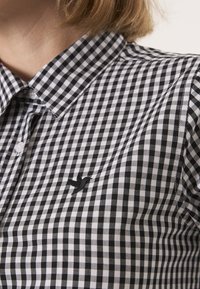 Camicia a quadri bianco e nero con colletto a punta, caratterizzata da un piccolo logo di uccello ricamato in nero sul petto.