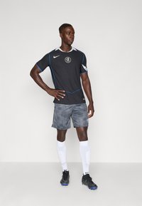 Maglia atletica nera con accenti blu e logo, abbinata a pantaloni corti grigi a fantasia, calze bianche e scarpe da calcio nere.