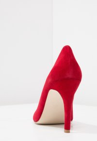 Röd mocka högklackad sko med spetsig tå och smal stilettklack. Slät textur och minimalistisk design.
