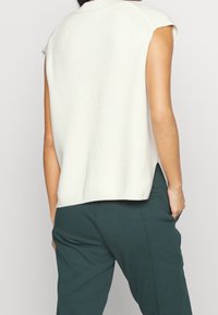 Top de punto sin mangas en crema claro, con textura acanalada, cuello redondo y aberturas laterales. Combinado con pantalones verde oscuro.