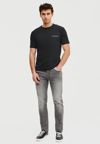 Czarny t-shirt z krótkim rękawem z białym logo, w połączeniu z szarymi dżinsami i czarnymi trampkami z białymi podeszwami. Model stoi na jednolitym tle.