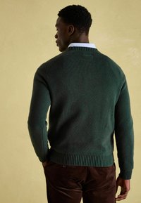 Mann, der mit dem Rücken zur Kamera steht, trägt einen dunkelgrünen Strickpullover über einem weißen Hemd mit Kragen und braunen Cordhosen vor einem beigen Hintergrund.