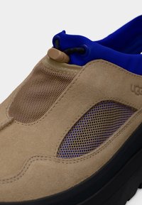 Μπεζ παπούτσι από σουέτ και δίχτυ, χωρίς κορδόνια, με μπλε νεοπρενένιο γιακά και μαύρη σόλα, με ρυθμιζόμενο κορδόνι κοντά στον αστράγαλο.