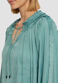 Helles Teal-Blusenshirt mit einem plissierten Design, Rüschenausschnitt und seitlichen Details. Verfügt über einen Bindeverschluss mit zwei dekorativen Schnurende.