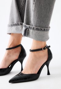 Tacones negros de punta con textura de cocodrilo, correa en el tobillo con herrajes dorados y un tacón alto delgado, combinados con unos jeans gris recortados.