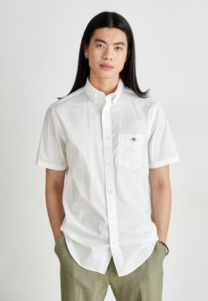 White short-sleeve button-up shirt with a chest pocket. Soft fabric, fitted design, and curved hemline. Simple branding on the pocket.
