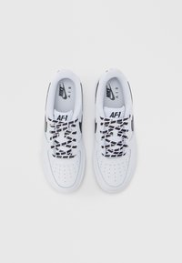 Λευκά sneakers Nike AF-1 με λείο δερμάτινο επάνω μέρος, διάτρητη μύτη, μαύρα λογότυπα και μαύρα κορδόνια με μοτίβο που φέρουν το κείμενο "NIKE".