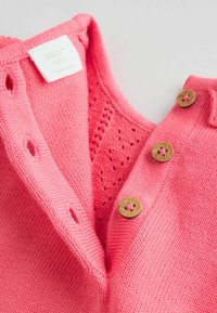 Rózsaszín kötött baba kardigán textúrázott mintával, három díszgombbal és "NEXT baby" feliratozású címkével a nyakán.