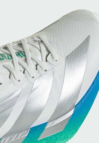 Close-up van een witte sportschoen met zilveren details, witte veters en een blauw-groene verloopzool.