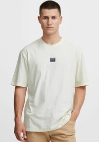 Lys gul kortærmet T-shirt med rund halsudsk�æring og et lille sort rektangulært logo-patch på brystet. Glat bomuldsstruktur.