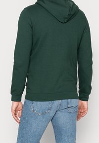 Dark green hoodie with a drawstring hood, ribbed cuffs, and hem. Worn with light blue jeans featuring a classic 5-pocket design.