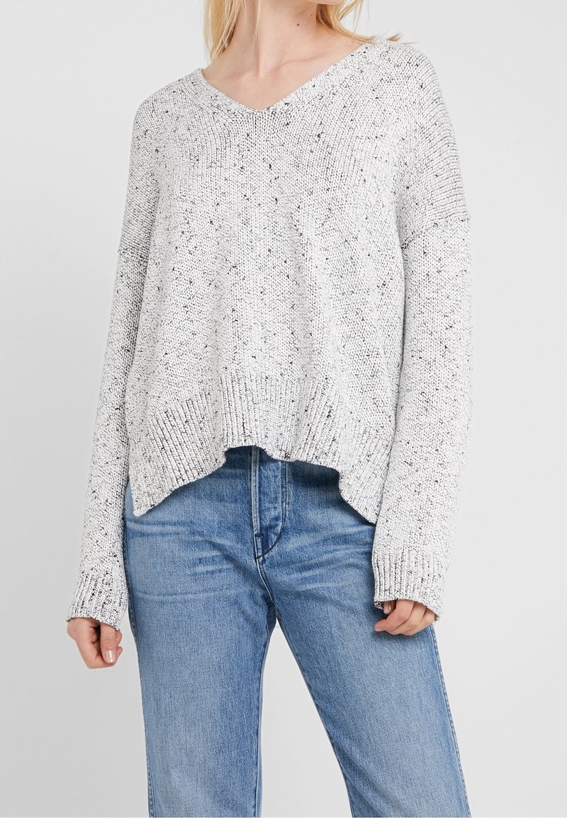 Pull en maille gris clair moucheté avec col en V et manches longues porté avec un jean bleu sur une personne des épaules à mi-cuisse.