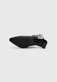Chaussure à talon haut noire avec un bout pointu, en matériau verni brillant et une semelle en caoutchouc texturé. Présente un lacet décoratif avec des accents de logo.