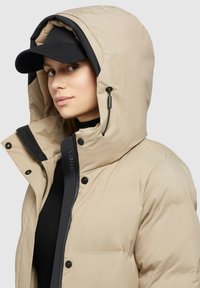 En kvinde iført en beige hættepufferjakke over en sort top og en sort kasket, der kigger lidt til siden mod en ensfarvet lys baggrund.
