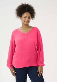 Blouse rose à manches longues avec un décolleté en V et un tissu légèrement texturé, présentant un détail de bouton au niveau du col et une coupe décontractée.