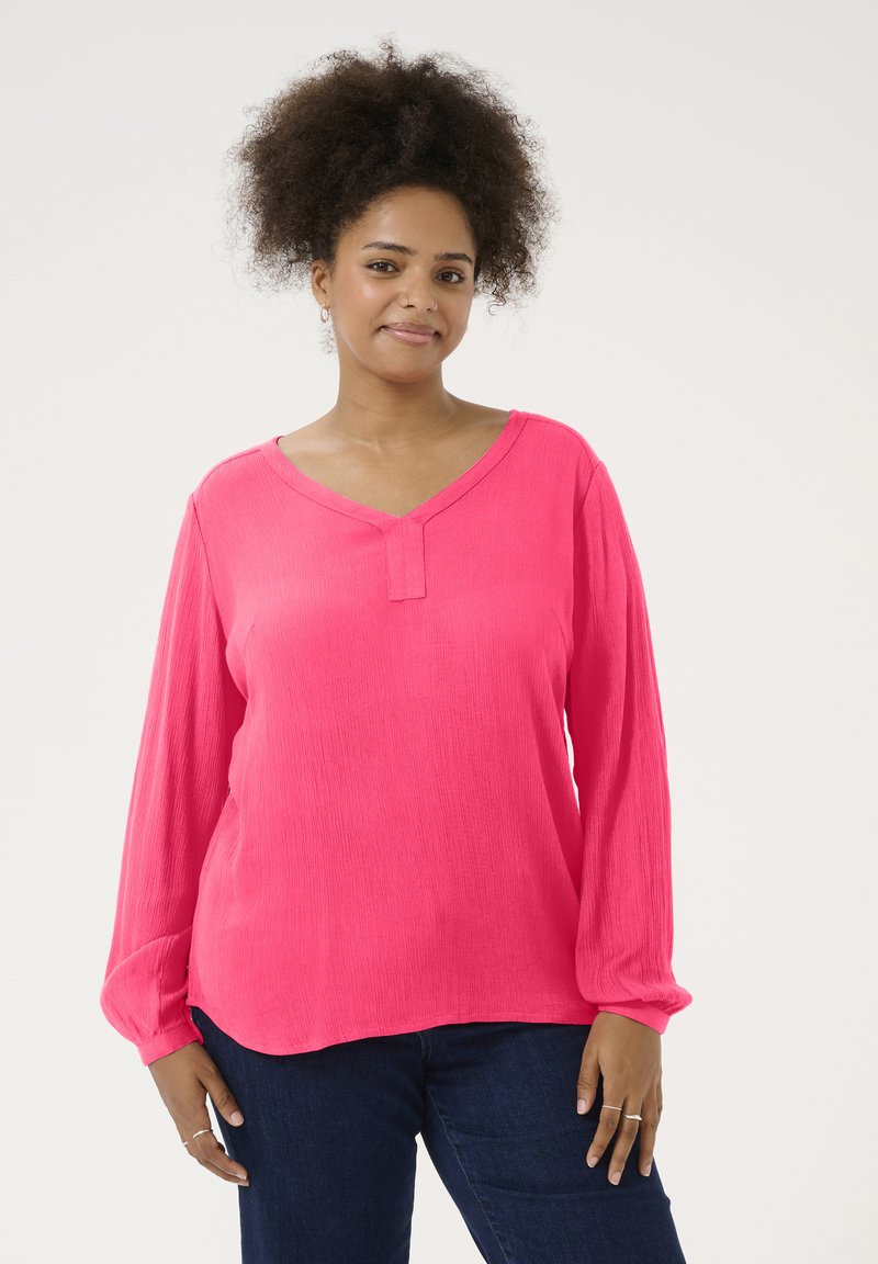Blouse rose à manches longues avec un décolleté en V et un tissu légèrement texturé, présentant un détail de bouton au niveau du col et une coupe décontractée.