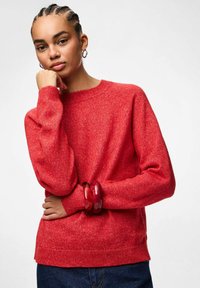 Raudonas megztinis su apvaliu iškirpimu, raukšlėtomis rankovėmis ir apačioje, derinamas su raudona apvalia apyranke. Fonas neutralus ir paprastas.