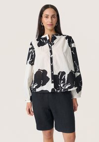 Langärmliges Hemd mit einem schwarz-weißen Blumenmuster, Knopfleiste vorne und elastischen Manschetten, kombiniert mit dunklen Shorts.