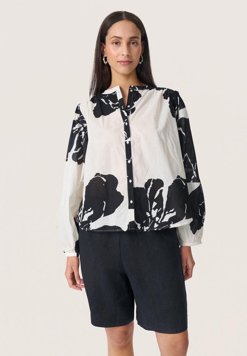 Langärmliges Hemd mit einem schwarz-weißen Blumenmuster, Knopfleiste vorne und elastischen Manschetten, kombiniert mit dunklen Shorts.