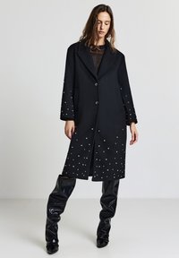 Cappotto di lana nero con un design sartoriale, colletto a punte, due bottoni e decorazioni in perline bianche attorno alle maniche inferiori e all'orlo.