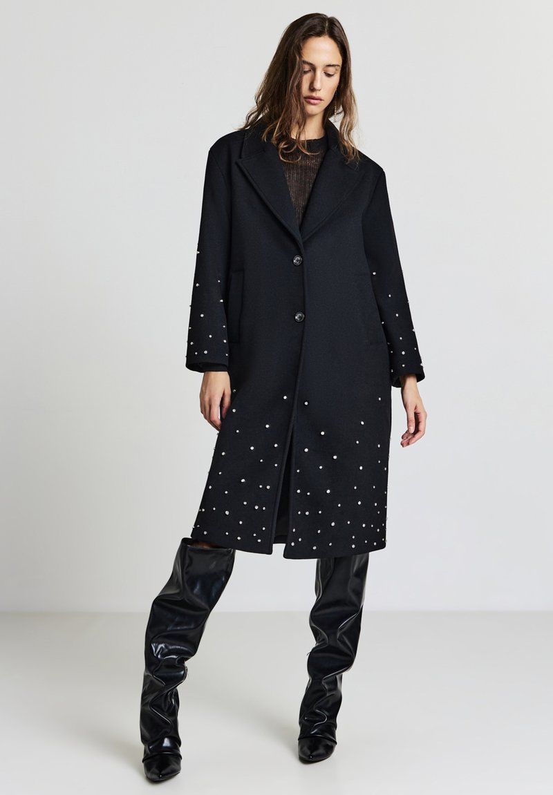 Cappotto di lana nero con un design sartoriale, colletto a punte, due bottoni e decorazioni in perline bianche attorno alle maniche inferiori e all'orlo.