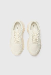 Baskets beige clair avec une tige en mesh texturé, des lacets plats et une semelle épaisse ; logo visible sur la doublure intérieure.