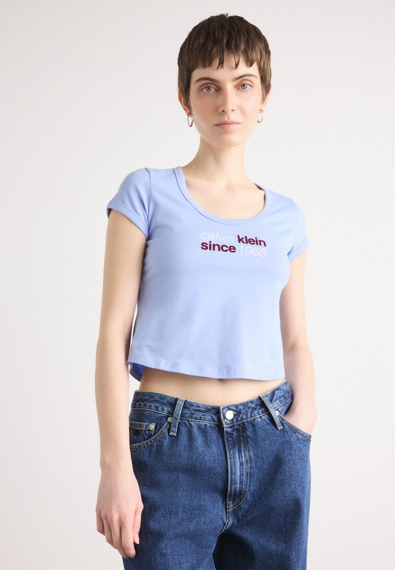 Light blue cropped t-shirt with short sleeves, featuring a red and black text logo "Calvin Klein since 1968". Paired with blue jeans.