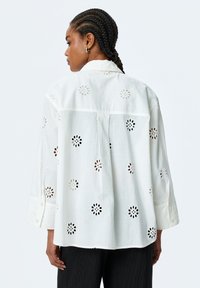 White shirt featuring floral cut-out patterns, button cuffs, and a relaxed fit. Made of lightweight fabric with subtle texture.