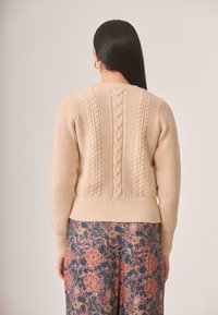 Suéter beige de punto con diseño de trenzas en la espalda, puños y dobladillo acanalados. Llevado sobre una falda con estampado floral que presenta colores vibrantes.