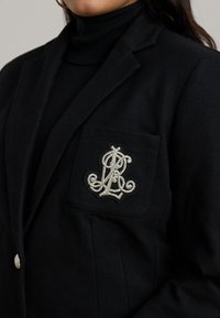Fekete blézer texturált anyagból, zsebén bonyolult fehér hímzett monogrammal, ezüst gombokkal kiegészítve.