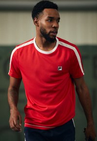 Camiseta deportiva roja con franjas blancas en los hombros, cuello redondo, logotipo en el pecho y mangas cortas, hecha de material texturizado y transpirable.