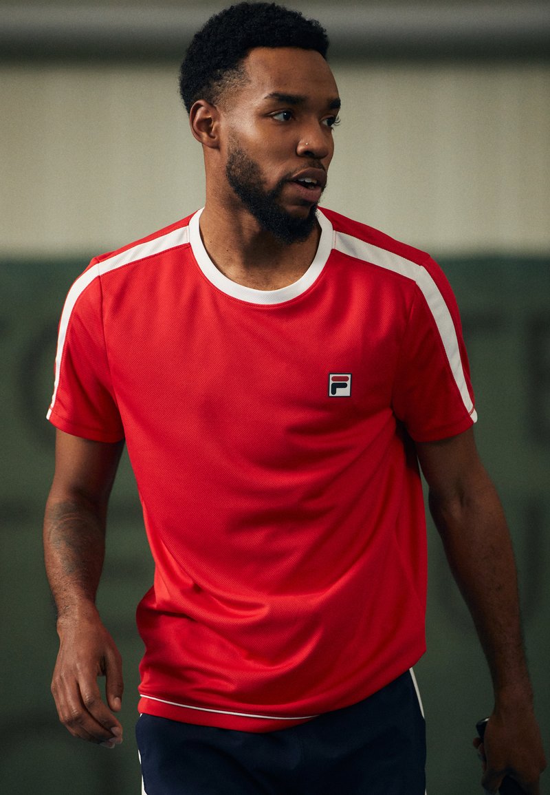 Camiseta deportiva roja con franjas blancas en los hombros, cuello redondo, logotipo en el pecho y mangas cortas, hecha de material texturizado y transpirable.