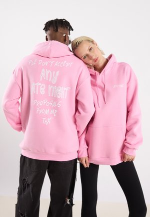 Les sweats à capuche roses présentent un texte blanc dans le dos, avec un grand message et des détails plus petits. Le tissu semble doux ; le design est surdimensionné avec une poche avant.