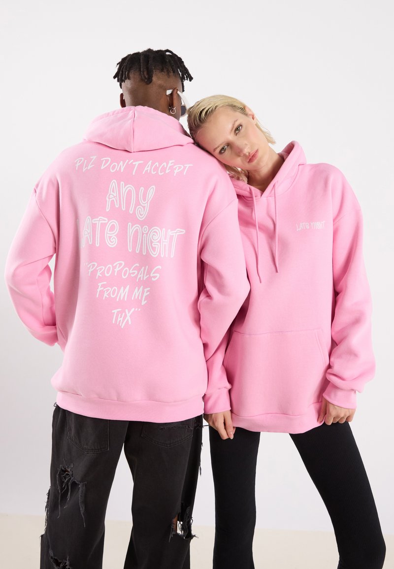 Le felpe rosa presentano un testo bianco sulla parte posteriore, con un grande messaggio e dettagli più piccoli. Il tessuto sembra morbido; il design è oversize e include una tasca frontale.